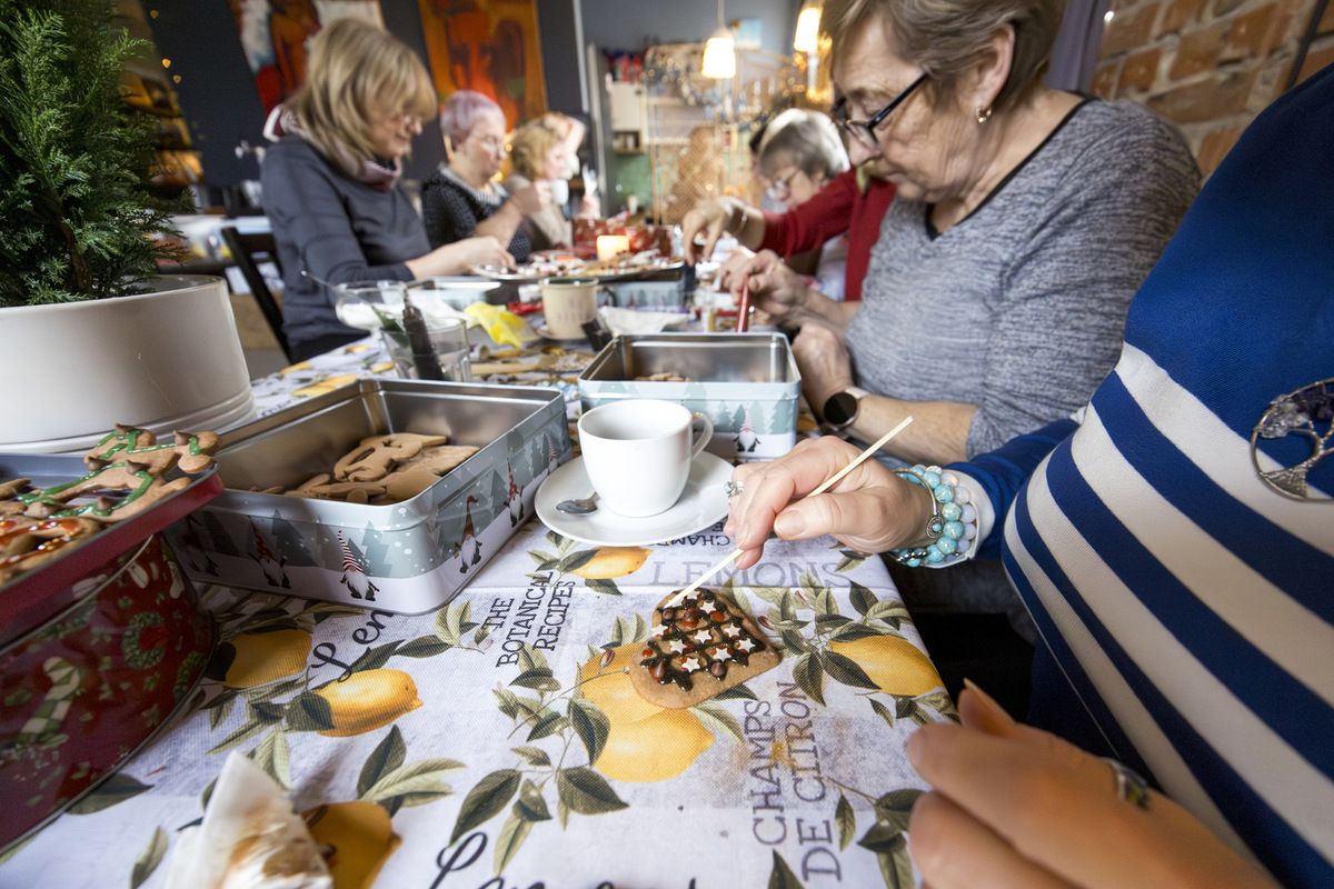 Kobiety dekorują pierniki przy długim stole podczas warsztatów „Oddech dla Mam”. Na pierwszym planie puszki z wypiekami i biała filiżanka. W tle uczestniczki pracują nad dekoracjami.